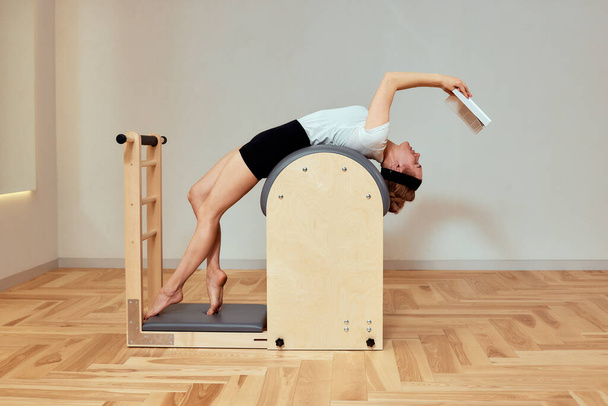woman doing pilates exercises on reformer, woman reading a book during pilates exercises on reformer, exercises on reformer to restore musculoskeletal system, pilates, reformerbed in a gym in a health - Foto, afbeelding