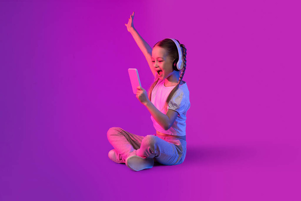 Ενθουσιασμένη ευτυχισμένη όμορφη preteen ξανθιά κοπέλα χρησιμοποιώντας σύγχρονα ασύρματα ακουστικά και smartphone σε πολύχρωμο φόντο στο φως νέον, κοιτάζοντας την οθόνη του κινητού τηλεφώνου και gesturing, τον έλεγχο νεότερο κινητό app - Φωτογραφία, εικόνα