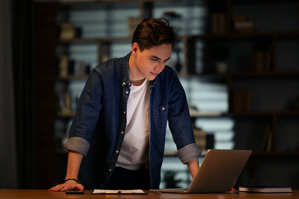 Positive handsome young man wearing casual outfit manager standing by workdesk at dark office, looking at laptop screen and smiling, employee have deadline, working late, copy space - Photo, Image