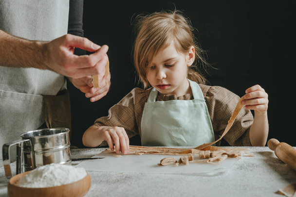 Unrecognizable father and daughter in aprons holding dough cut into strips on a white table against a dark wall. Parent with child preparing homemade pastries or noodles. Selective focus. - Fotoğraf, Görsel
