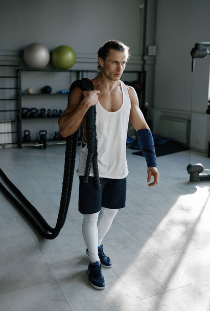 Jongeman met een sterke lichaamsvorm die fitnesstouwen op de schouders draagt en tegen de achtergrond van een moderne sporthal loopt - Foto, afbeelding