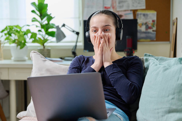 Surprised emotional joyful teenage girl in headphones looking at laptop screen while lying on couch at home. Amazement, surprise, astonishment concept, internet online video technology - Valokuva, kuva