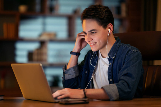 Fiatal barátságos jóképű férfi alkalmi ruha projekt menedzser van online találkozó késő este, ül íróasztal egyedül éjszaka sötét irodában, a modern PC laptop és fülhallgató, másolás hely - Fotó, kép