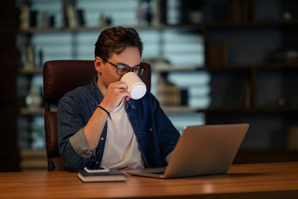 Ciężko pracujący młody pracownik pracujący do późna w biurze, facet ubrany w strój służbowy i okulary siedzący przy biurku w ciemnym pokoju, korzystający z nowoczesnego laptopa, pijący kawę, kopiujący przestrzeń - Zdjęcie, obraz