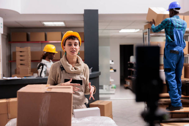 Storehouse worker recording smartphone video with inventory management explanation for shipping company trainee. Smiling young woman in warehouse overall and helmet talking at mobile phone camera - Photo, Image