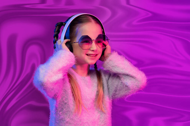 Retrato de dulce linda niña rubia linda en edad escolar en gafas de sol con auriculares inalámbricos modernos, niño feliz escuchando música, divertirse sobre fondo de lámina olográfica iridiscente rosa - Foto, imagen