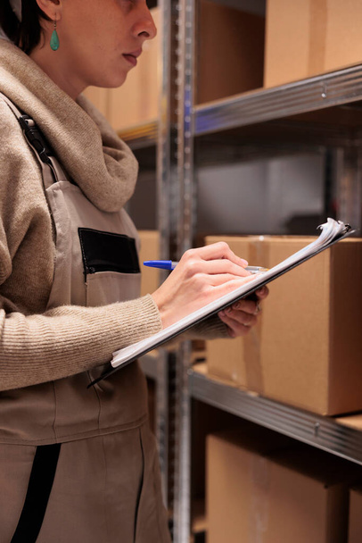 Storehouse employee checking goods in stock and writing on clipboard. Warehouse supervisor wearing unifrom jumpsuit verifying freight in stockroom, marking checklist close up - Fotó, kép