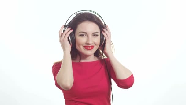 Beautiful model in red swaying along with the music in big headphones in slow motion isolated on white background medium shot - Footage, Video
