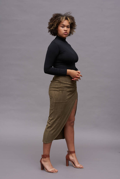Mulher intercultural elegante jovem em gola alta preta, saia marrom longa e sandálias de salto alto posando contra fundo cinza - Foto, Imagem