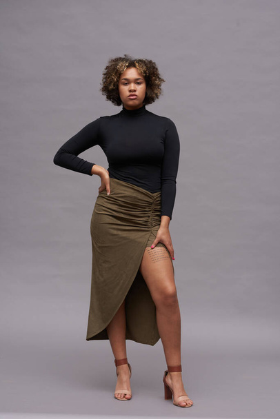 Young African American well-dressed woman in turtleneck and long skirt looking at camera while standing with right hand on her waist - Фото, изображение