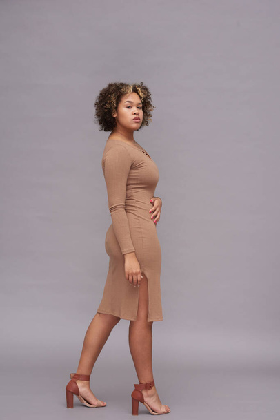 Young African American woman in casual dress of beige color looking at you while posing in front of camera against grey wall in studio - Foto, Bild