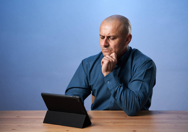 Mature business man sitting at his desk with his tablet, on blue background - Foto, Imagen