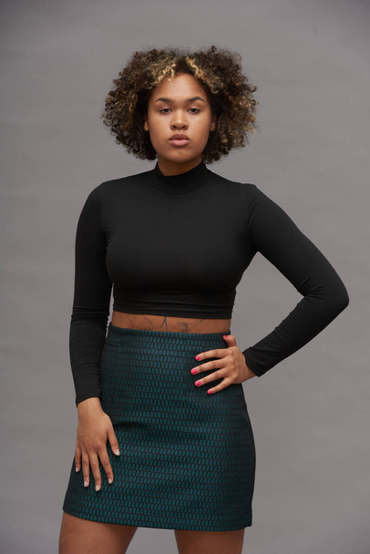 Young elegant woman in cropped top and skirt posing in front of camera in studio while standing in isolation against grey background - Photo, Image