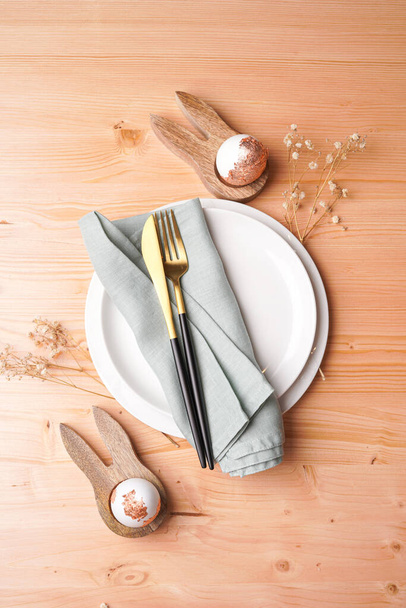 Μια κορυφαία άποψη σε ένα άδειο λευκό κεραμικό πιάτο, χρυσά μαχαιροπήρουνα σε λινό χαρτοπετσέτα, κουνελάκια σε σχήμα θήκες αυγών, χρυσά foiled αυγά σε ένα ξύλινο φόντο, αντίγραφο χώρου, πάνω άποψη - Φωτογραφία, εικόνα