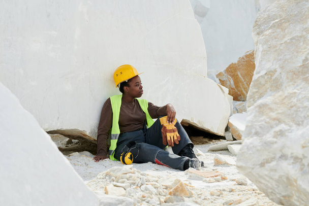 Young restful female worker in uniform and safety helmet having break after work while sitting against huge marble rock at quarry - Photo, Image