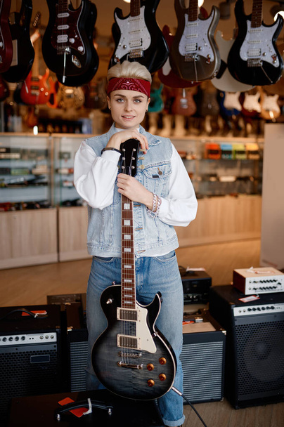 Ritratto di giovane musicista hipster donna con chitarra elettrica e sorriso sul viso guardando la macchina fotografica in piedi sopra negozio di musica professionale - Foto, immagini