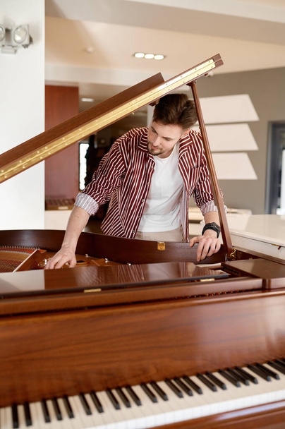 Music instrument shop store concept. Young man buyer looking on tuning keys inside grand piano under lid - Photo, Image