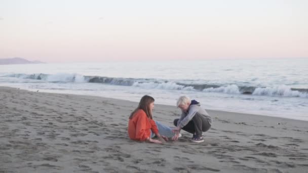 Een paar tieners, een jongen en een meisje, zitten op een zandstrand aan zee en praten. - Video