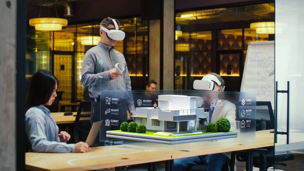 Male designer uses VR headset and wireless controllers, presents smart home exterior design project in virtual reality to coworkers. Modern hi-tech company. 3D hologram. Future digital technologies. - 写真・画像