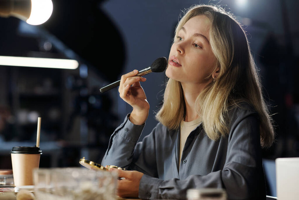 Young blond woman with cosmetic brush applying powder on her face while sitting by workplace and preparing for shooting - Fotó, kép