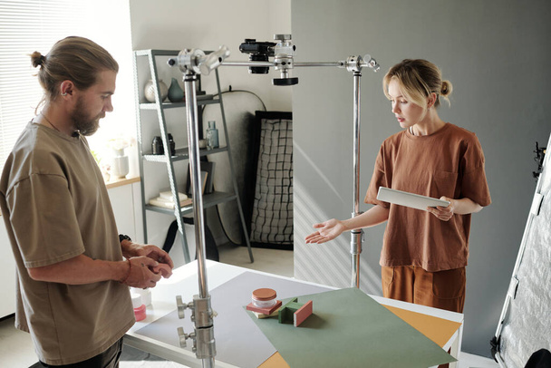 Jovem com tablet apontando para a composição com objetos na mesa enquanto a apresenta ao fotógrafo antes de fotografar - Foto, Imagem