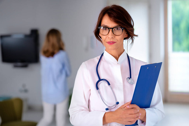 Vrouwelijke arts draagt labjas en stethoscoop en houdt klembord in haar handen terwijl ze in de gang van het ziekenhuis staat - Foto, afbeelding