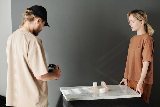 Jongeman met fotocamera maakt foto 's van compositie op marmeren platform terwijl zijn assistent voor hem staat - Foto, afbeelding