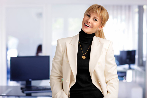 Close-up of businesswoman smiling and looking at camera. Blond haired female wearing white blazer while standing at the office. Copy space.  - Photo, Image