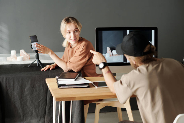 Jeune assistant regardant photographe pointant vers smartphone avec image à l'écran lors de la discussion de nouvelles photos en studio - Photo, image