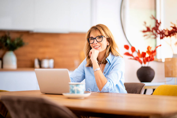 Middle aged woman sitting at her dining room table and using earphones and laptop while having video call. Home office.  - 写真・画像