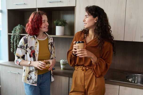 Két fiatal, elegáns női alkalmazott, intelligens alkalmi ruhákban kávéznak és ebédelnek, miközben a konyhában állnak és szünetben beszélgetnek. - Fotó, kép