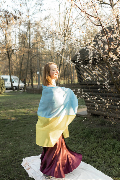 Жінка в сукні з прапором Украніана біля дерев у весняному парку на тлі квітучого дерева сакури.. - Фото, зображення