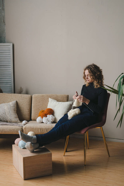 Woman knitting with croche, sitting on the armchair near sofa using knitting yarn. Crochet thick threads. Home comfort. - Fotó, kép