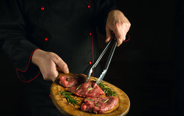 Professiona chef holds a cutting board with raw beef steaks before barbecue. Preparation for roasting meat with rosemary and spices. Black space for restaurant recipe or hotel menu - Photo, image