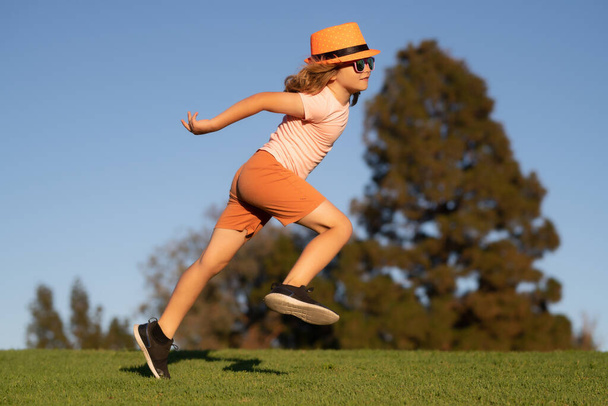 Kid boy running on green grass near blue sky in summer park. Little runners outdoors in summer nature. Sporty child running in a park. Outdoor sports and fitness for children - 写真・画像