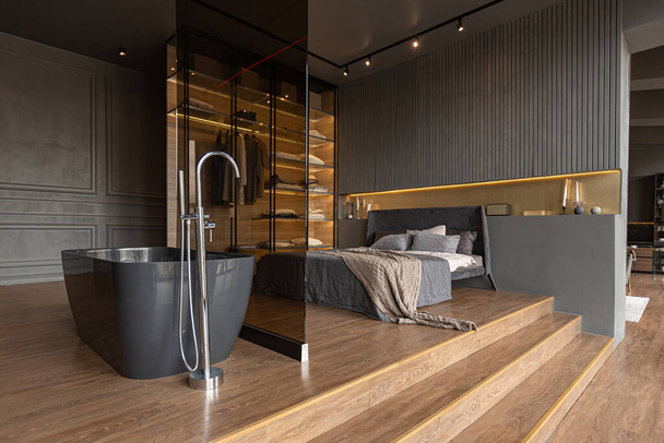 bedroom and freestanding bath behind a glass partition in a chic expensive interior of a luxury home with a dark modern design with wood trim and led light - Photo, Image