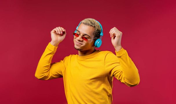 Positive american man listening music, enjoying dance with headphones on red studio background. Radio, wireless modern sound technology, online player. High quality photo - Fotografie, Obrázek
