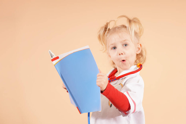 A little girl with a stethoscope holds a book in her hands and looks into it. - Foto, Imagen