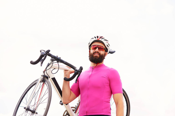 ヘルメットと眼鏡のピンクのジャケットを着た男性のサイクリストは、空に向かって肩に自転車を持ち、自転車で練習し、自転車で街を歩いています。. - 写真・画像