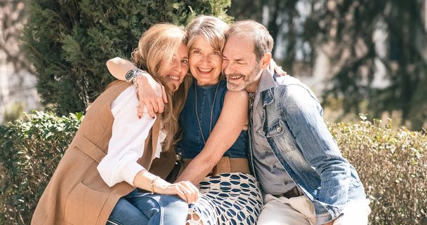 Two senior women and one senior man are sitting in a sunny park, smiling and hugging each other. Friendship, love, and support in the golden years - Photo, Image