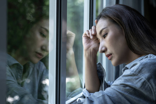 Portrait of sad and worried Asian woman looking through the window at home - Photo, Image