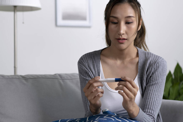 Junge Asiatin hält Schwangerschaftstest nach Hause und sieht im Wohnzimmer glücklich aus - Foto, Bild