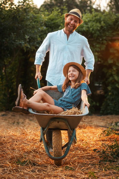 Χαρούμενος γενειοφόρος με ψάθινο καπέλο σπρώχνει ένα καρότσι με ένα χαρούμενο κοριτσάκι. Πατέρας και κόρη απολαμβάνουν το καλοκαίρι μαζί στην πίσω αυλή. Κάθετα. Η έννοια της κηπουρικής. - Φωτογραφία, εικόνα
