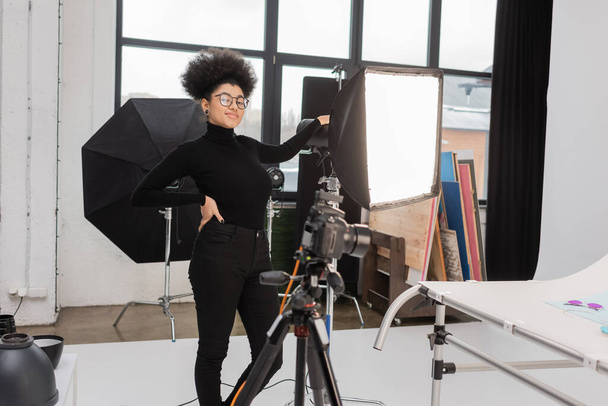 elégedett afro-amerikai tartalomkészítő álló kézzel csípőn közel softbox reflektor és elmosódott digitális fényképezőgép a fotó stúdióban - Fotó, kép