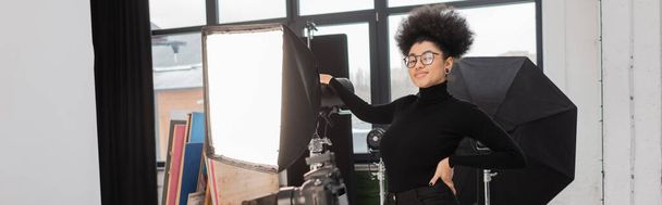 heureux producteur de contenu afro-américain posant avec la main sur la hanche près du réflecteur softbox dans le studio photo, bannière - Photo, image