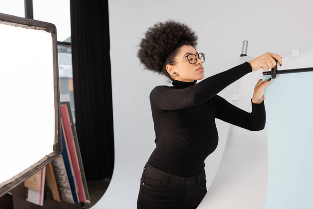 productor de contenido afroamericano en anteojos instalando hoja de fondo cerca del reflector en estudio fotográfico - Foto, imagen