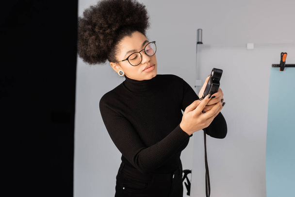 elegante gerente de contenido afroamericano en cuello alto negro mirando el medidor de exposición en el estudio de fotos - Foto, Imagen