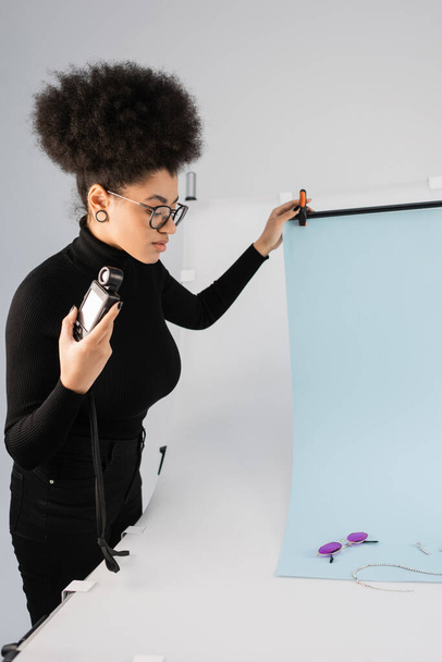 Afro-Amerikan içerik üreticisi beyaz arka plan örtüsünün yanında duruyor ve çekim masasında son moda güneş gözlükleri var. - Fotoğraf, Görsel