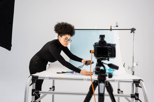 Fabricante de contenido afroamericano mirando a la cámara digital borrosa mientras sostiene el medidor de exposición cerca de la mesa de tiro en el moderno estudio de fotografía - Foto, imagen