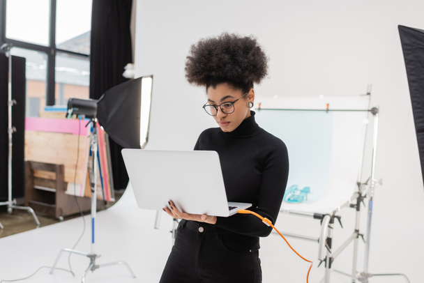 africký americký obsah výrobce v brýlích při pohledu na notebook v blízkosti softbox reflektor a fotografický stůl ve fotostudiu - Fotografie, Obrázek